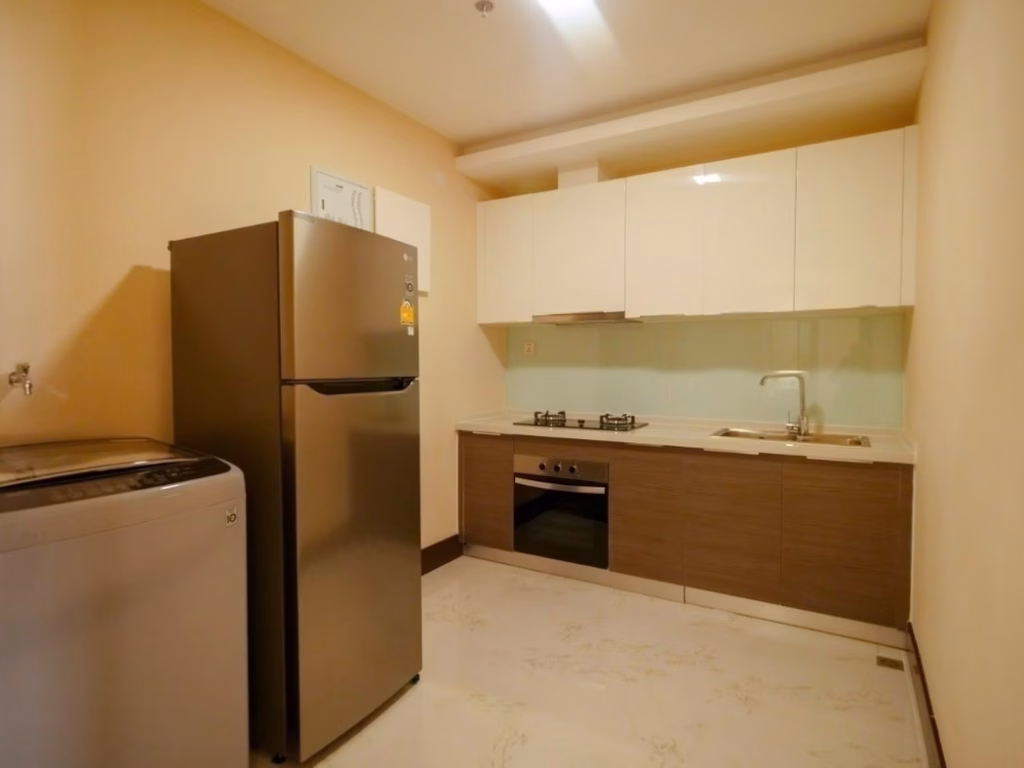 Modern kitchen in a 2 bedroom condo with beige walls, white cabinets, a stainless steel refrigerator, a washing machine, built-in oven, gas stove, and sink on a long countertop with a light-colored floor.