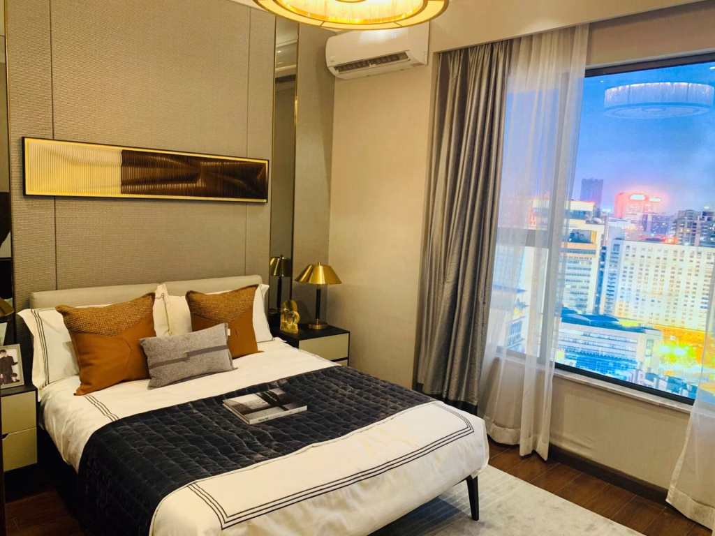 Modern bedroom at Agile Sky Residence with a neatly made bed, decorative pillows, gold lamps on nightstands, abstract wall art above the bed, and a large window overlooking the city skyline at dusk.