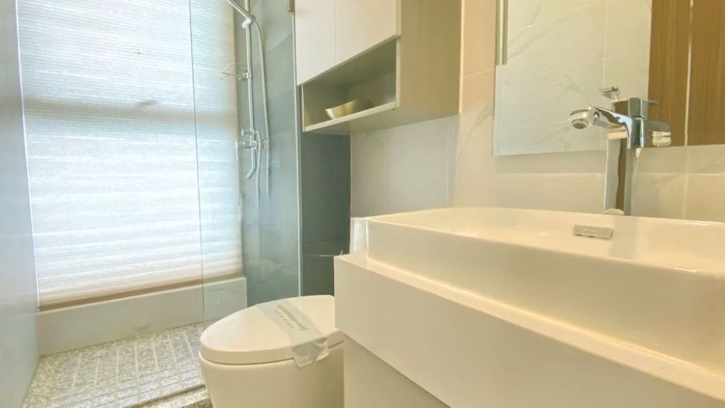 A modern bathroom by Morgan EnMaison Studios features a white sink, chrome faucet, toilet, and glass shower enclosure. Light-colored tiles and a large frosted window fill the space with natural light.