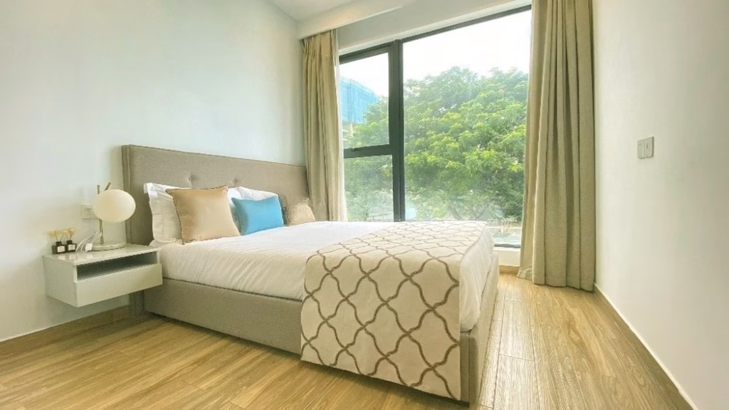 A modern bedroom by Morgan EnMaison Studios features a large bed with light-colored bedding, a side table with a lamp, wood flooring, beige curtains, and a large window framing lush greenery outside.
