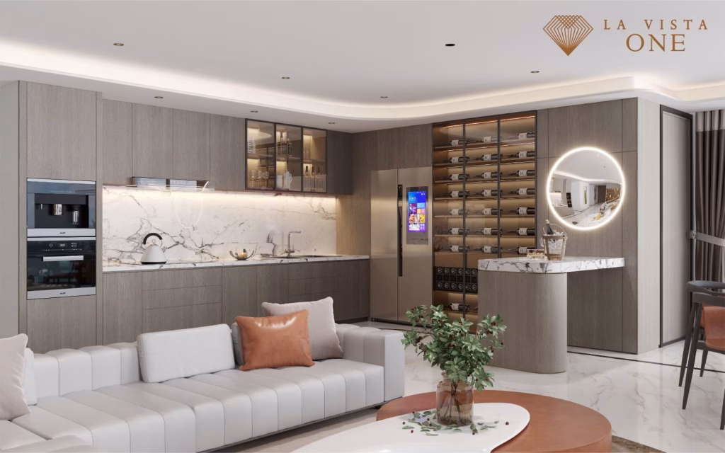 Modern kitchen and living room in a luxury penthouse with sleek wood cabinets, marble countertops, smart refrigerator, wine rack, and circular mirror. A white sofa with brown cushions sits in the foreground. La Vista One logo is top right.
