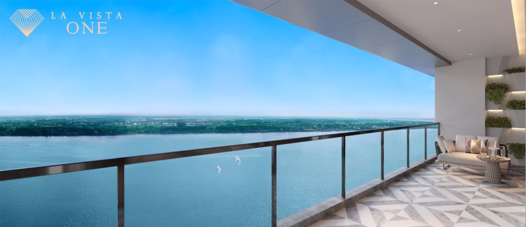 A modern balcony in a luxury penthouse with a glass railing overlooks a wide river and distant green landscape; there is outdoor seating with cushions and a small table. The sky is clear and bright with La Vista One text in the corner.