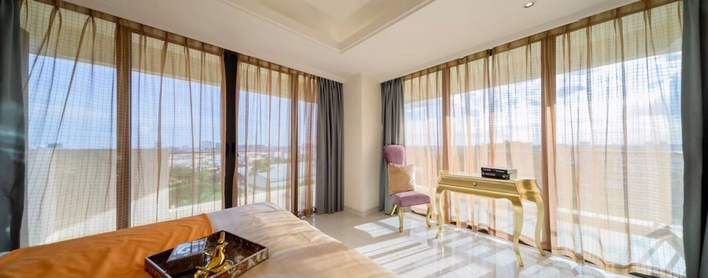 A bright Star City bedroom with large floor-to-ceiling windows draped in sheer beige curtains, featuring a golden desk, a purple chair, and part of a bed in the foreground as sunlight streams into the room.