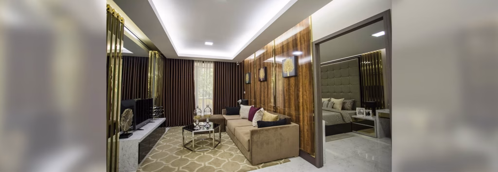 Modern living room at Orkide The Royal featuring a beige sofa, glass coffee table, wall-mounted TV, patterned rug, wooden accent wall, and large windows with dark curtains; bedroom visible through an open doorway.