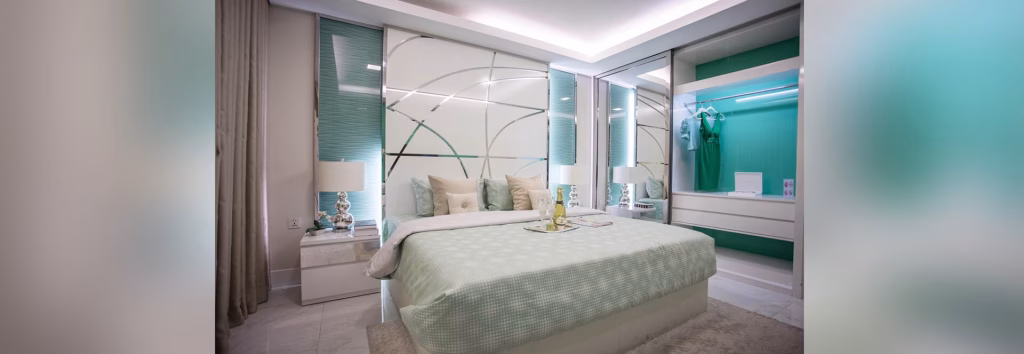 A modern bedroom with a large bed, light green bedding, two bedside tables with lamps, mirrored walls, and a built-in closet featuring a teal dress and shelves. The room exudes elegant decor and soft lighting inspired by Orkide The Royal.