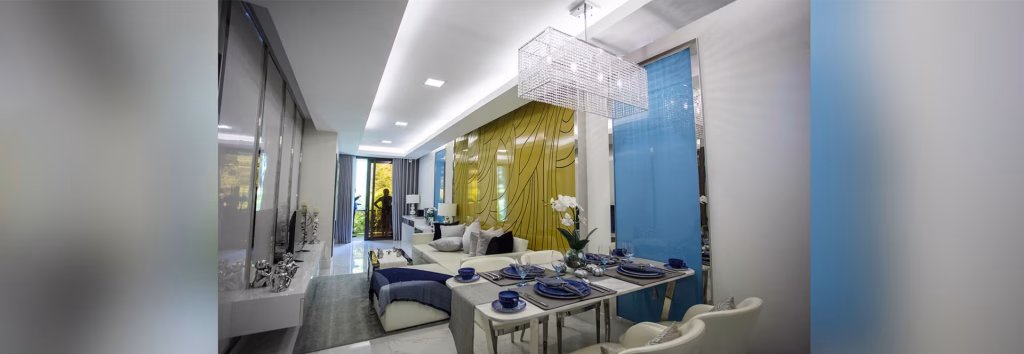 Modern living and dining area at Orkide The Royal with a glass dining table set for four, white chairs, beige sofa, yellow accent wall, blue decor, and large windows letting in natural light. Contemporary lighting overhead.