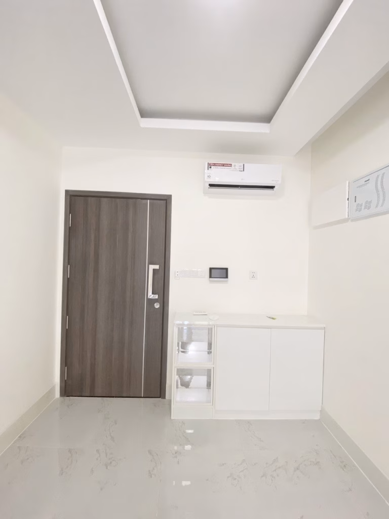 A modern, minimalistic 1 bedroom condo featuring a wooden door, a white air conditioner, a sleek storage cabinet, a small clear drawer unit, and light-colored tiled floors. The walls and ceiling are plain white for a clean look.