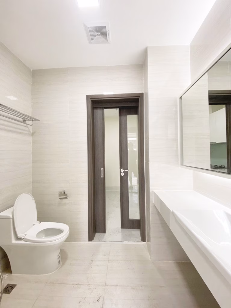 A modern, minimalistic bathroom in a 1 bedroom condo features light-colored tiles, a white toilet, a wall-mounted towel rack, a rectangular sink, a large mirror, and a dark wooden door with glass panels.