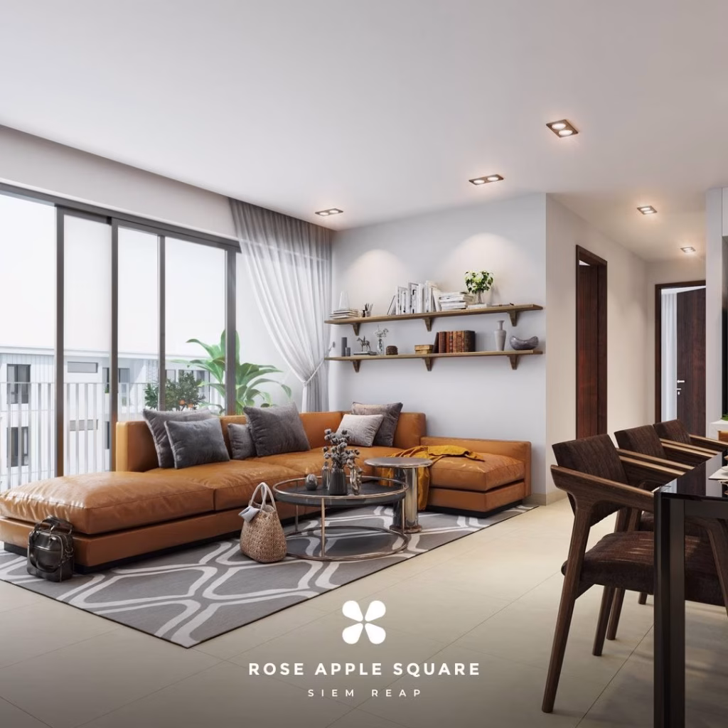 A modern living room at Rose Apple Square features a brown sectional sofa, gray and white rug, coffee table, bookshelves, and large windows for natural light. The stylish, inviting space flows seamlessly into the dining area on the right.