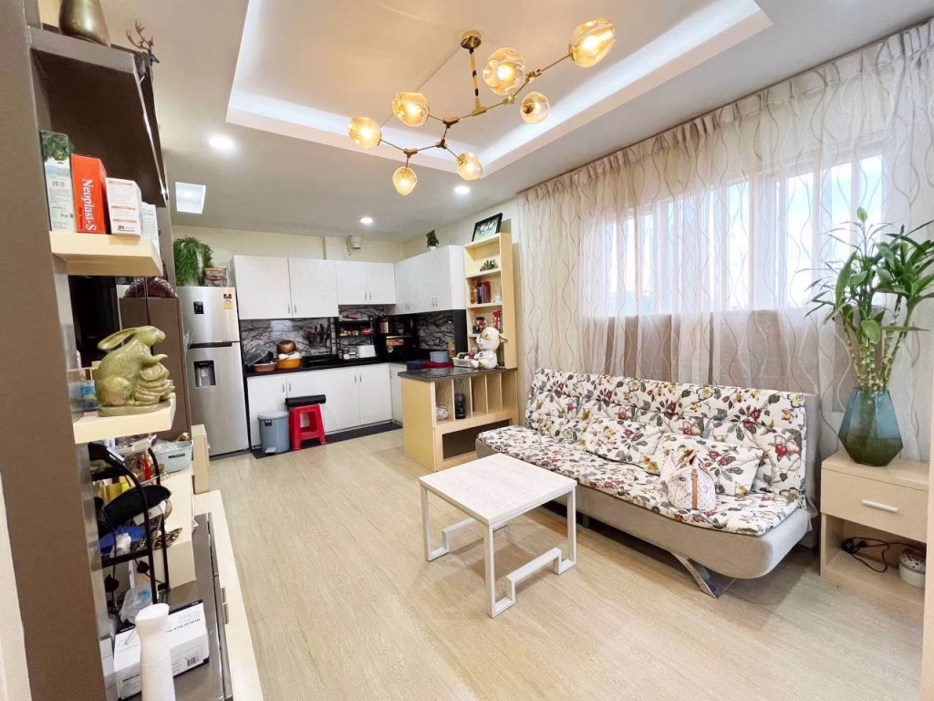 A bright, modern 2 bedroom condo living room with a floral sofa, white coffee table, shelves, potted plants, and an open kitchen in the background. Ceiling lights and large windows with sheer curtains provide ample light.