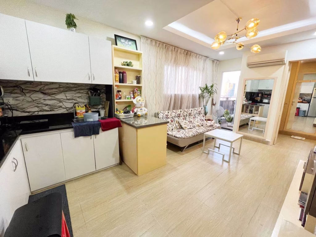 A bright, modern 2 bedroom condo living room features a small kitchen area with white cabinets, a beige island, floral sofa, coffee table, shelves with decor, large windows with sheer curtains, and a ceiling light fixture.