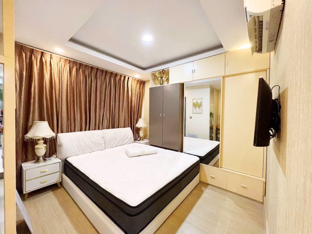A modern 2 bedroom condo features a spacious bedroom with a large bed, white bedding, bedside tables with lamps, brown curtains, built-in mirrored wardrobes, and a wall-mounted TV. The well-lit room boasts a minimalist design for stylish comfort.