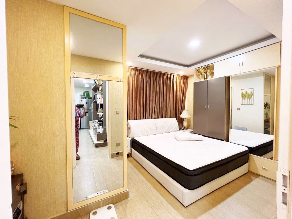 A modern 2 bedroom condo bedroom with a double bed, white and gray bedding, large wardrobe, floor-length mirror, bedside table with a lamp, brown curtains, and wooden flooring. A person is partially visible in the mirror.