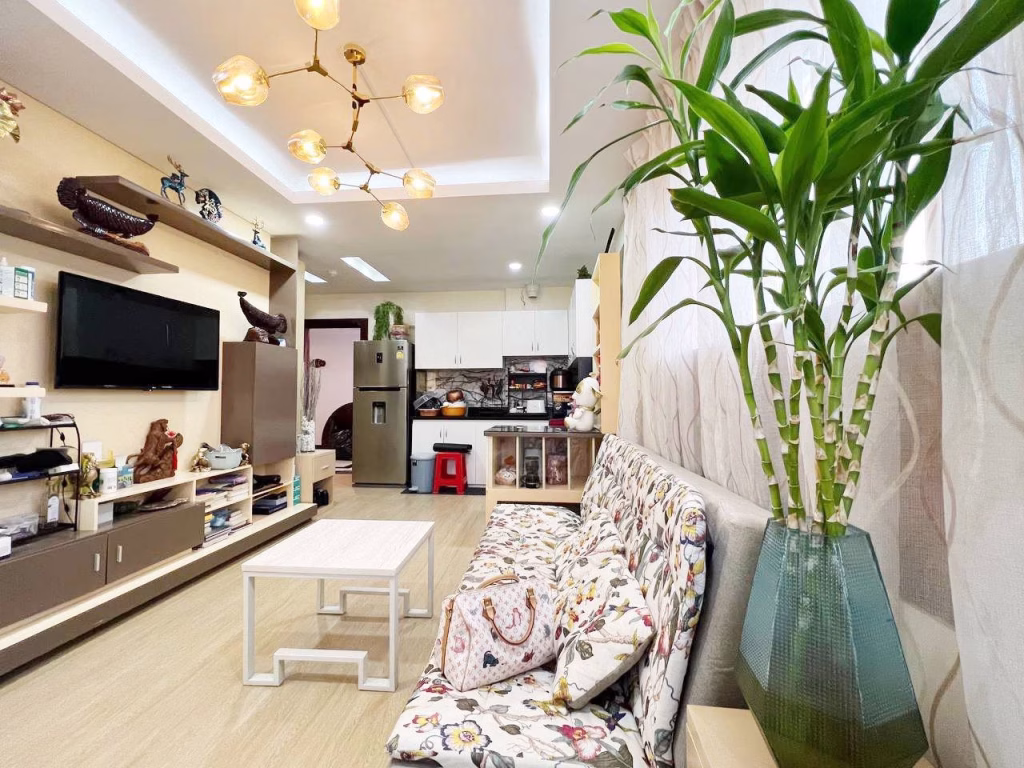 A bright, modern living room in a 2 bedroom condo features a floral-patterned sofa, glass vase with green plants, white coffee table, wall-mounted TV, shelves with decor, and an open view into the kitchen area with appliances and utensils.