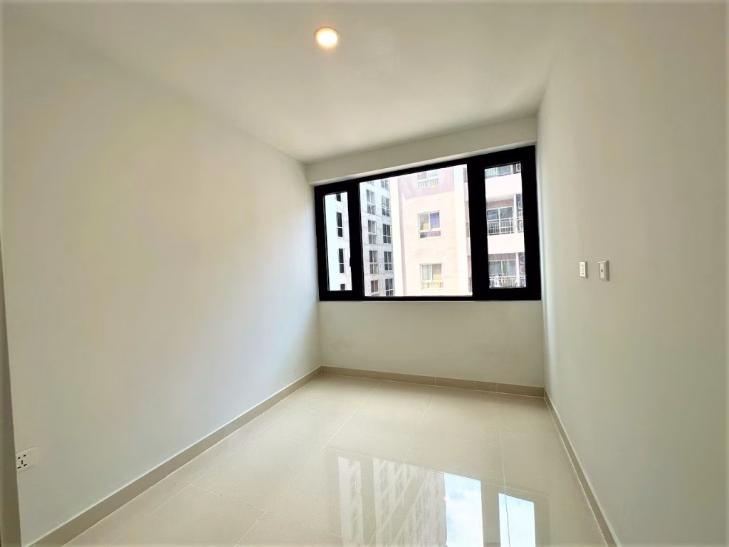 A small, empty room with white walls, a tiled floor, a ceiling light, and a large window that fills this 2 bedroom condo with natural light while offering views of tall buildings outside.