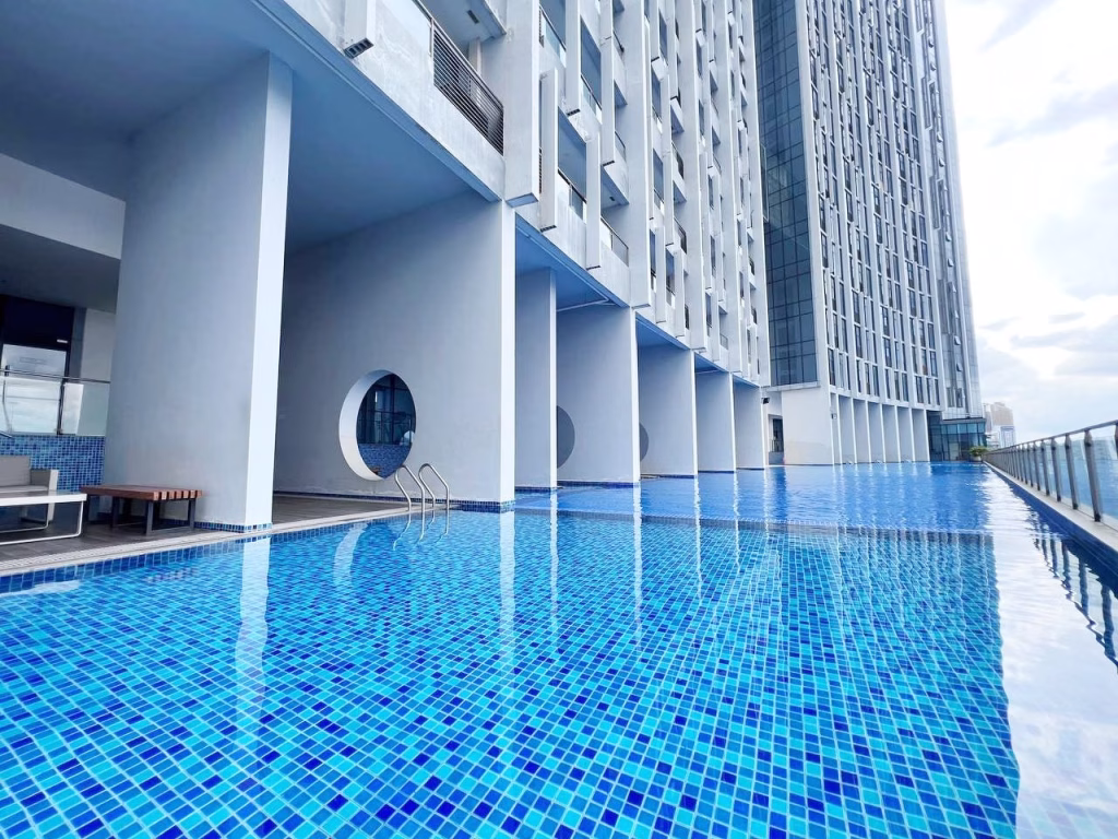 Modern outdoor swimming pool with blue mosaic tiles beside a tall, white 2 bedroom condo building featuring circular windows and glass balconies, under a bright sky.
