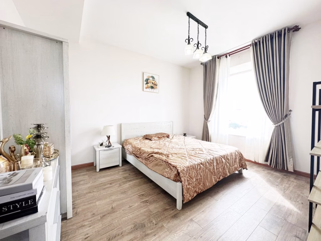 A bright, minimalist bedroom in a 3 bedroom apartment features a double bed with beige bedding, wooden floor, white furniture, a large window with gray curtains, and decorative items on shelves and the nightstand.
