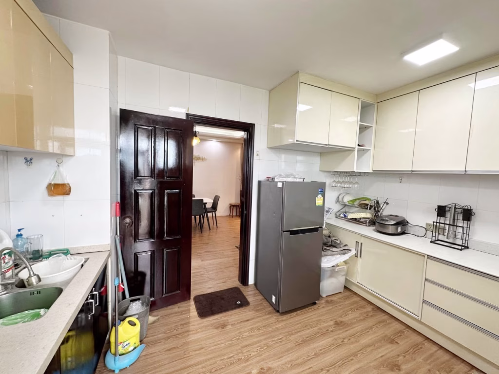 A modern kitchen in a 3 bedroom apartment features light cabinets, a silver refrigerator, countertops with appliances, a sink with cleaning supplies, and an open door leading to a dining area with a wooden floor.