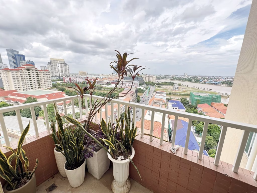 Three potted plants are arranged on the balcony of a 3 bedroom apartment with a white railing, overlooking a cityscape of buildings, roads, and greenery under a cloudy sky.