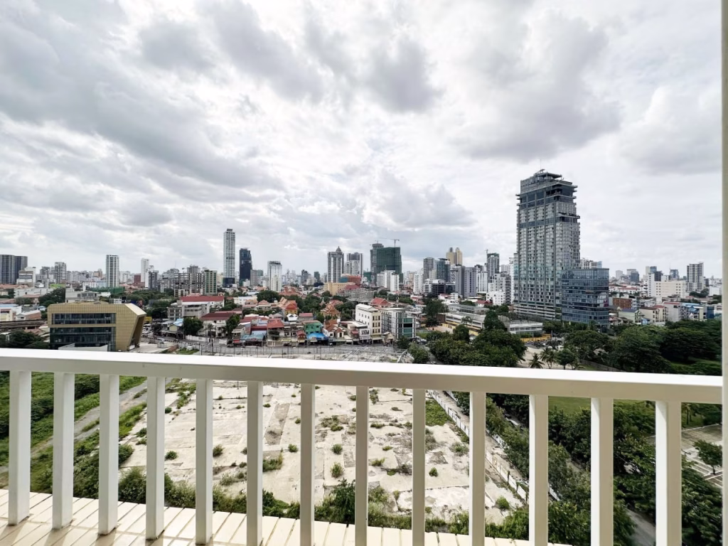 Cityscape view from a balcony of a 3 bedroom apartment with white railing, showing modern high-rise buildings, mid-rise structures, and a sandy, undeveloped lot below under a cloudy sky.