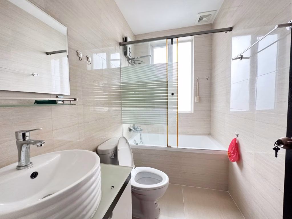 Modern bathroom in a 3 bedroom apartment with beige tiled walls, a white sink and toilet, a bathtub with glass shower screen, wall mirror, towel racks, and a window for natural light. A red plastic bag hangs near the toilet.