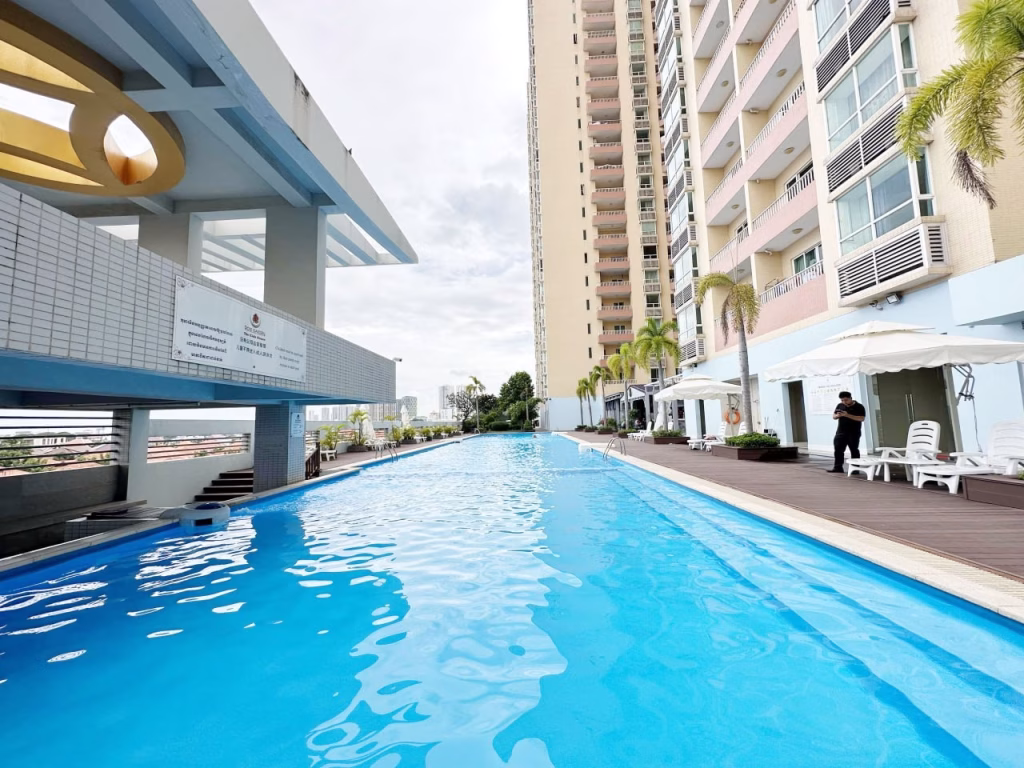 A long outdoor swimming pool stretches beside a tall, modern 3 bedroom apartment building with balconies. Lounge chairs, palm trees, and two people near white tents line the wooden deck under a cloudy sky.