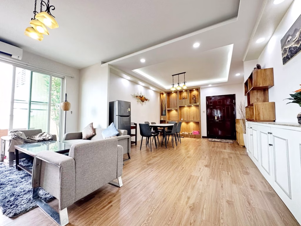 Modern open-plan living and dining room in a 3 bedroom apartment with wooden flooring, gray sofa set, glass coffee table, dining table with chairs, built-in shelves, and warm lighting. Large window lets in natural light.