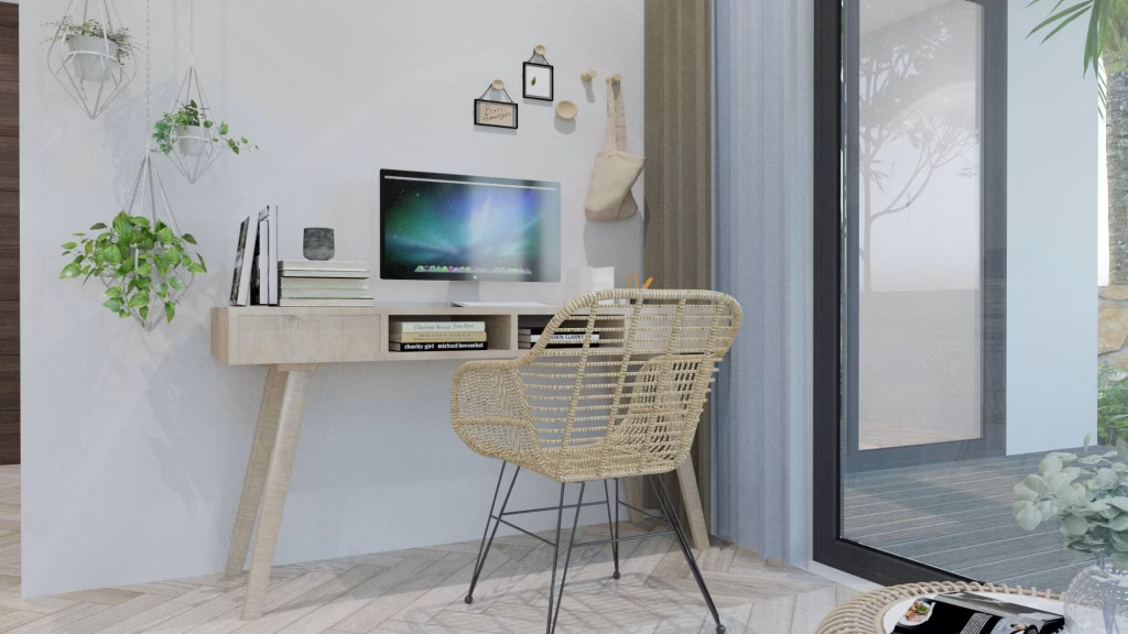 A modern home office with a wooden desk, computer, books, potted plants, and a woven chair. Bright natural light pours in through a large glass door, offering lush views—perfect for those seeking kampot homes for sale with inviting workspaces.