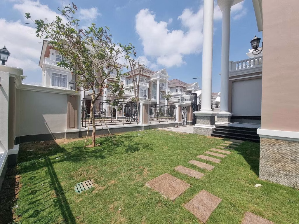 A sunny yard with a small tree, green lawn, and stone paths sits beside a beautiful corner villa with white columns and a black fence. Neighboring houses are visible in the background under a blue sky.