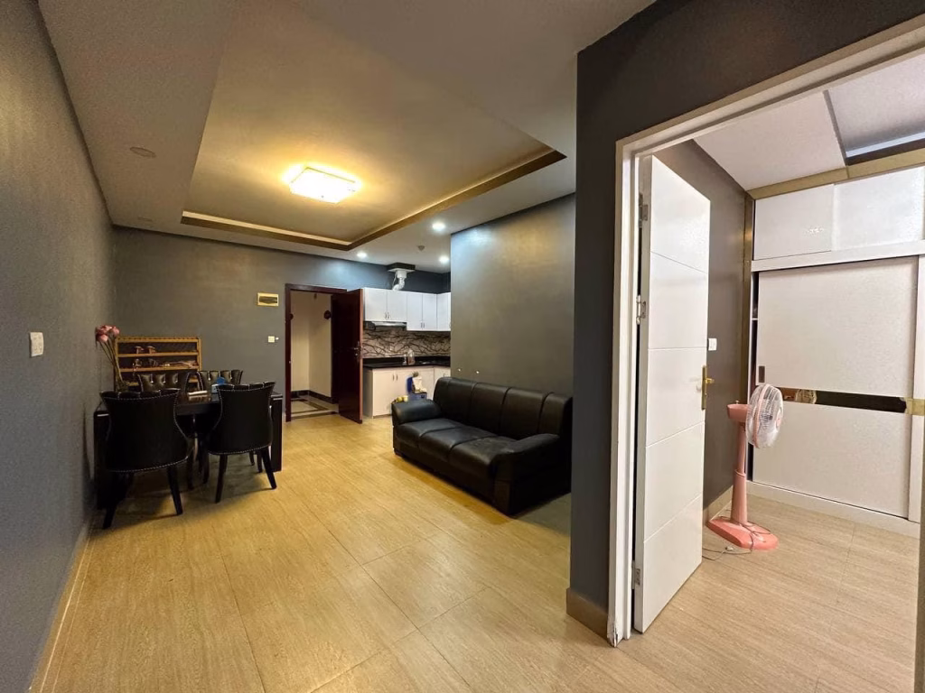 A modern condo for sale featuring a dining table and chairs, a black sofa, an open kitchen, and a doorway leading to a room with a pink standing fan and white sliding closet doors. The walls are painted gray.