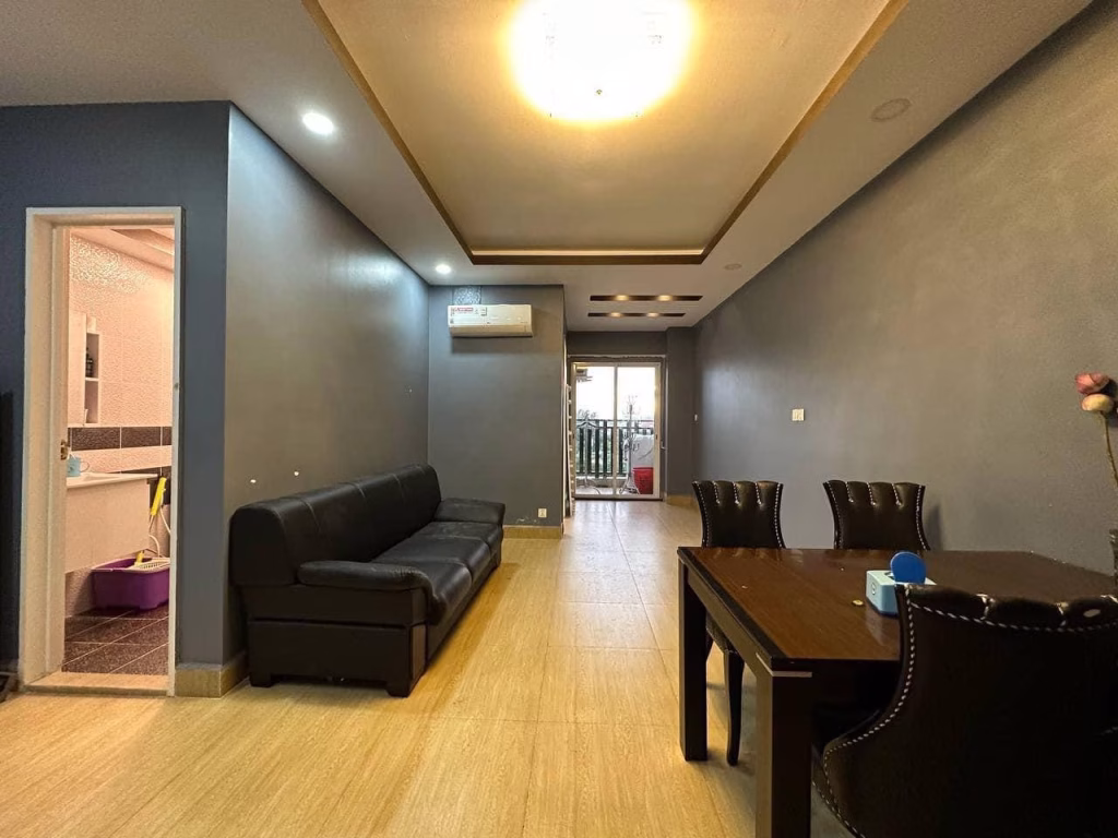 A modern condo for sale featuring a living and dining area with gray walls, a black sofa, wooden dining table, and ceiling light. An open door reveals the bathroom, while a balcony is visible at the far end.