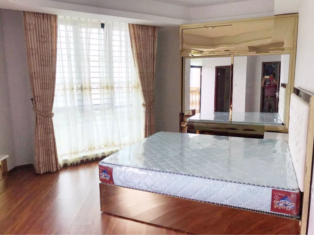 A bright condo for sale features a bedroom with large windows draped in sheer curtains, a bare mattress on a bed frame, wooden flooring, and a mirrored wardrobe reflecting part of the airy space.