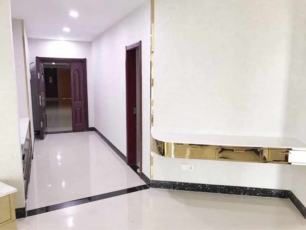 A clean, modern hallway in this condo for sale features white glossy floors, cream-colored walls, dark brown doors, and a gold-trimmed floating shelf. Ceiling lights illuminate the space, giving it a bright look.