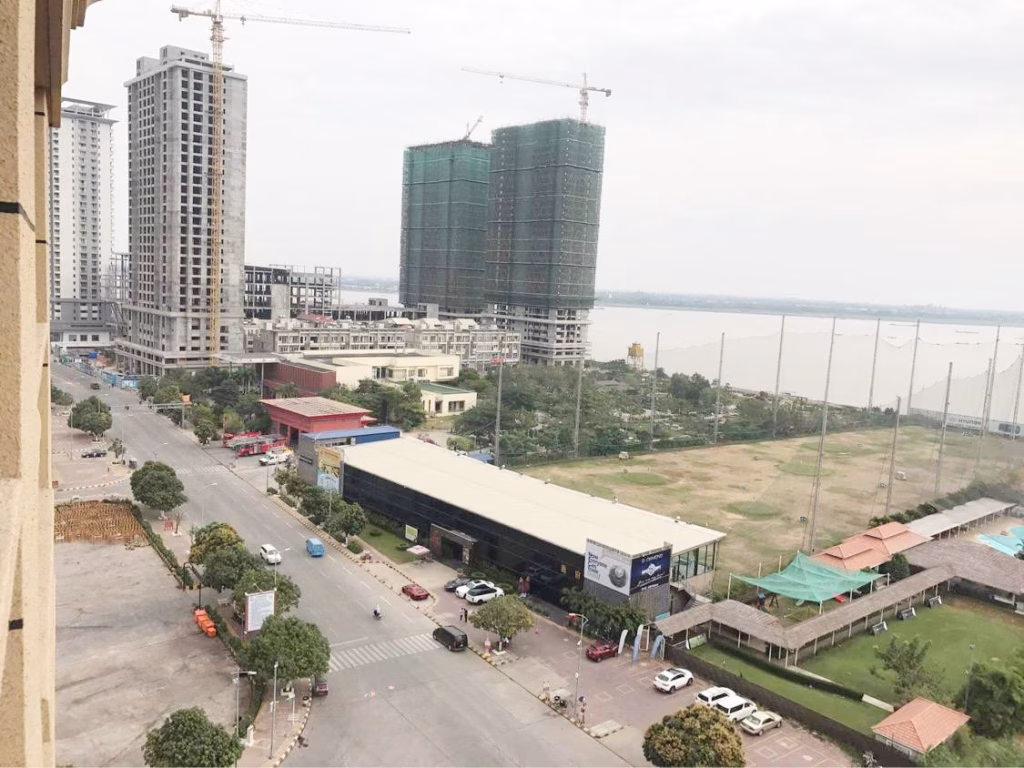 A cityscape view shows high-rise buildings under construction and a wide road with cars, while a condo for sale overlooks greenery, a river, and a sports facility with a netted area and swimming pool on the right under cloudy skies.