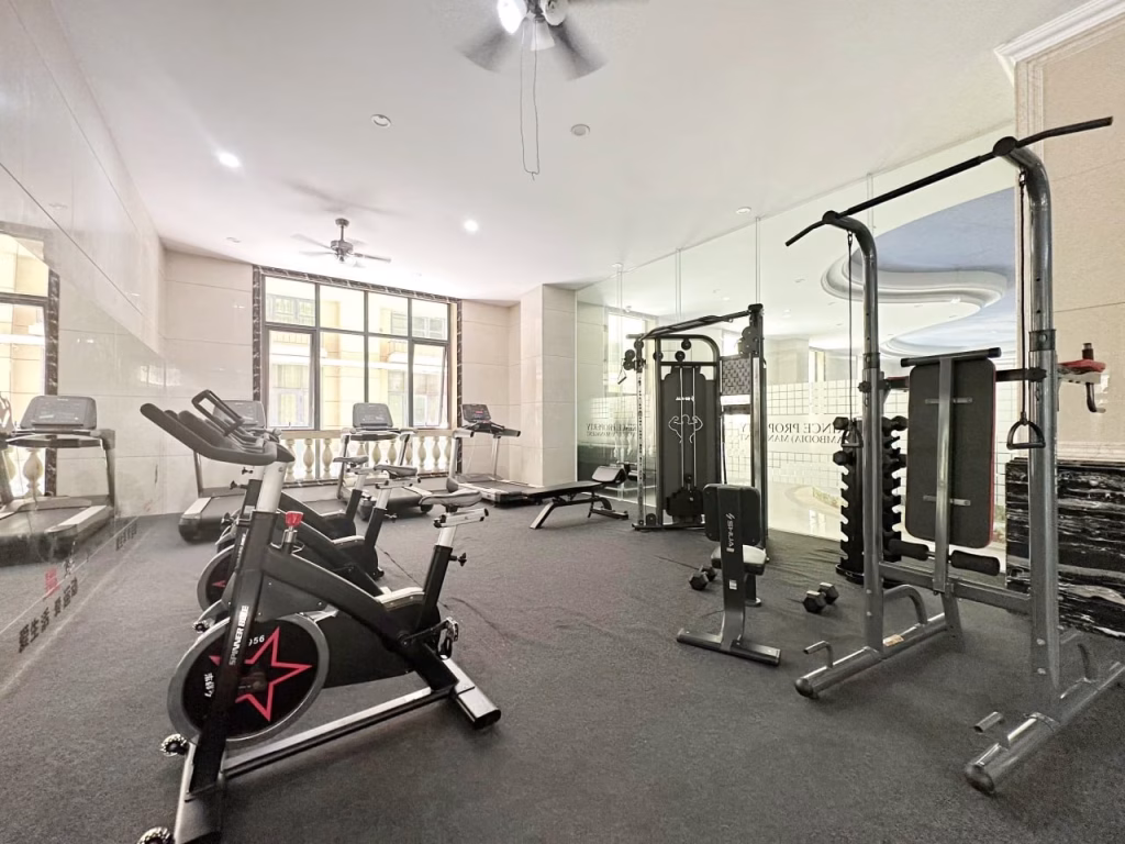 A modern gym room in this condo for sale features cardio machines like exercise bikes and treadmills, plus a cable machine, bench, and free weights. Large windows and mirrors create a bright, spacious atmosphere.