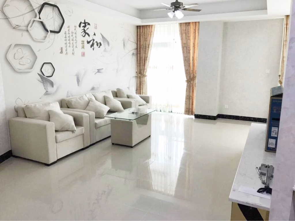 A modern, bright living room in this condo for sale features a white sofa set, glass coffee table, wall art with Chinese characters, light curtains, and large windows. The shiny floor beautifully reflects the stylish decor and natural light.