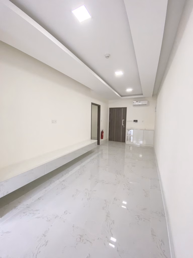A bright, modern hallway at Orkide The Royal with white marble floors, cream walls, a long bench on one side, a wooden door at the end, and recessed ceiling lights. An air conditioner and fire extinguisher are visible.