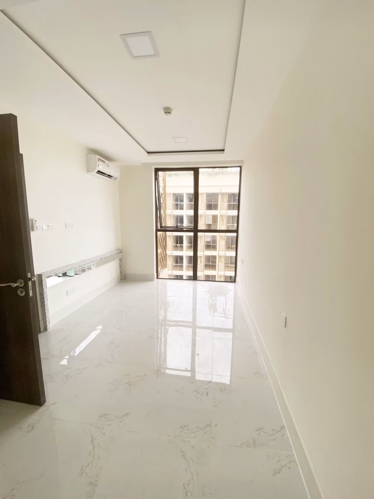A bright, empty room at Orkide The Royal with white marble flooring, cream-colored walls, a large window with a balcony view, an open wooden door, and a wall-mounted air conditioner.