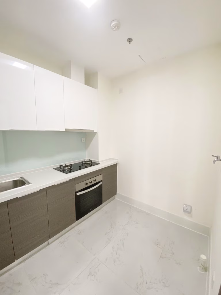 A modern, minimalist kitchen at Orkide The Royal features white upper cabinets, wood-toned lower cabinets, a built-in oven, gas stovetop, sink, and light marble-patterned floor tiles against plain, light-colored walls.