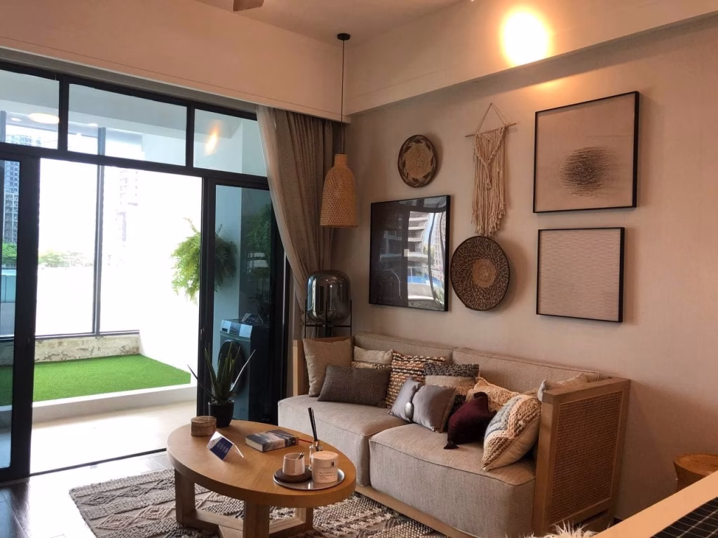 A cozy Sihanoukville studio for sale features a beige sofa, patterned pillows, and wall art. Large sliding glass doors open to a balcony with artificial grass and potted plants, filling the living room with warm, natural light.