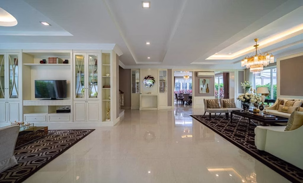 Spacious, elegant living room in a modern house for sale, featuring glossy tiled floors, built-in shelves, TV, comfortable seating, chandeliers, large windows, and a stylish view into the dining area.