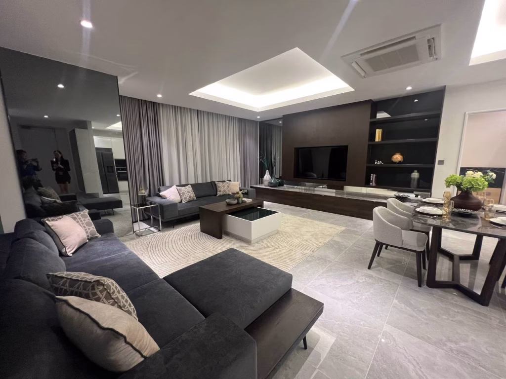 A modern 3 bedroom apartment living room with a large dark sectional sofa, neutral-toned pillows, a round rug, built-in shelving, wall-mounted TV, dining table for four, floor-to-ceiling curtains, and two people reflected in the mirror.