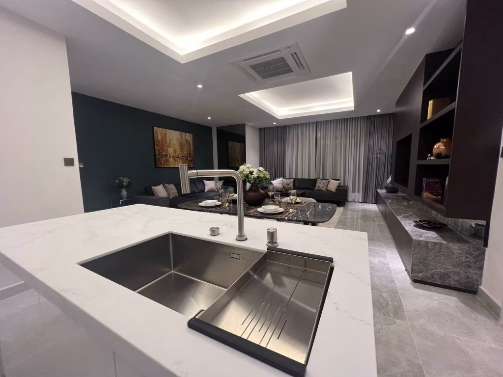 A modern open-concept living room in a stylish 3 bedroom apartment, featuring a marble-topped kitchen island and sink, a cozy seating area with sofa and cushions, coffee table with flowers, and a large TV on a sleek media unit.