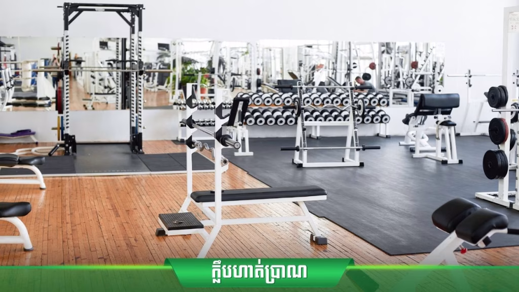A modern gym at Oasis Harmony villas offers various exercise equipment, including weight benches, dumbbells, and squat racks. The room features large mirrors, wooden flooring, and green and white text in Khmer at the bottom of the image.