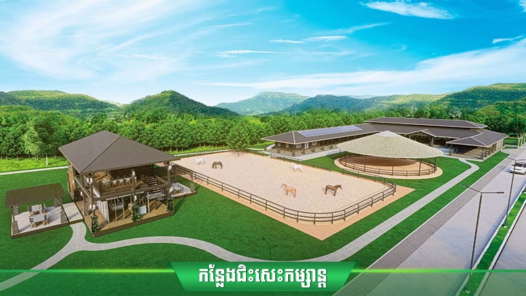 An aerial view of Oasis Harmony villas near a horse ranch with a large riding arena, grazing horses, stable buildings, lush green trees, and mountains in the background beneath a clear blue sky.
