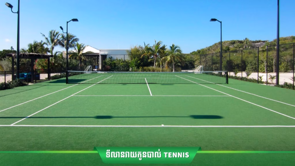 A green outdoor tennis court at Oasis Harmony Villas is surrounded by palm trees and lush greenery beneath a clear sky. The word TENNIS appears on a banner below in English and another script.