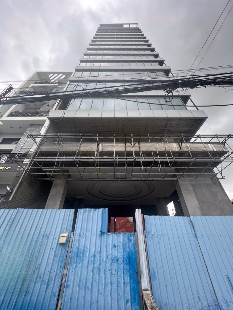 A tall prime office building under construction is viewed from below, with scaffolding on the lower floors and blue metal barriers in front. Overhead, power lines cross the cloudy sky.