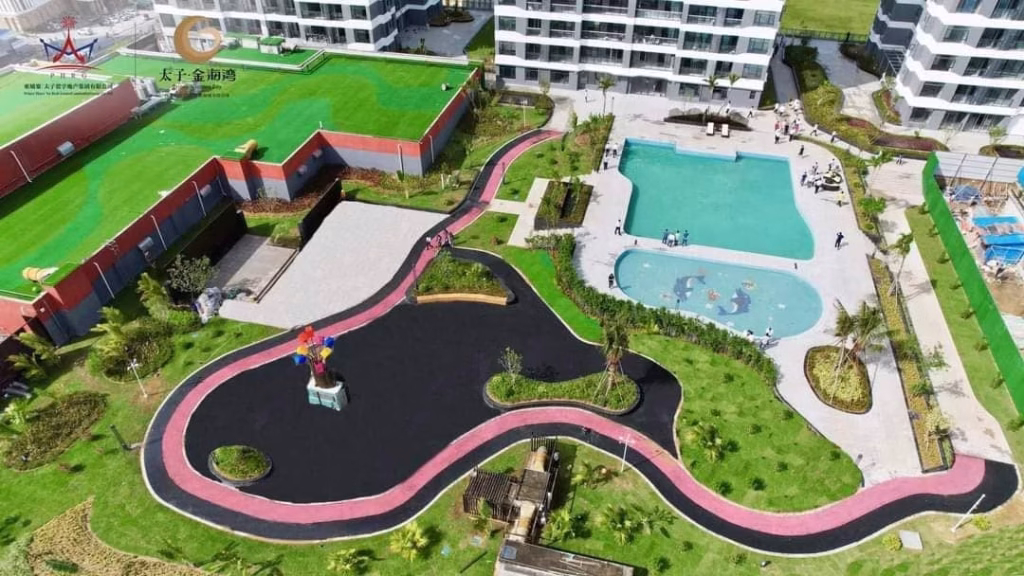 Aerial view of a modern residential complex in Sihanoukville with white buildings, a winding walking path, two swimming pools, green lawns, and landscaped gardens—ideal for those seeking a studio for sale.