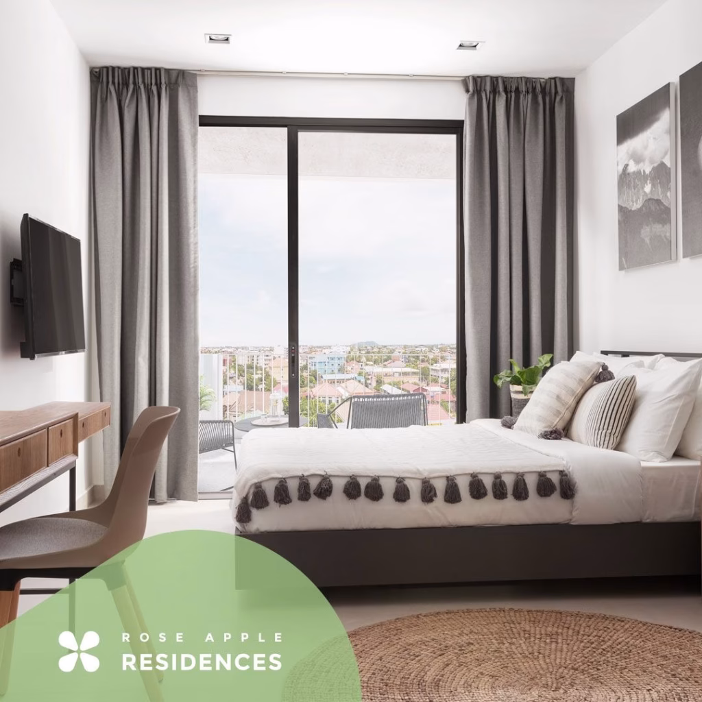 Modern bedroom at Rose Apple Square with a neatly made bed, desk, wall-mounted TV, and large glass doors leading to a balcony with a city view. Gray curtains frame the doors, while neutral decor creates a calm, inviting atmosphere.