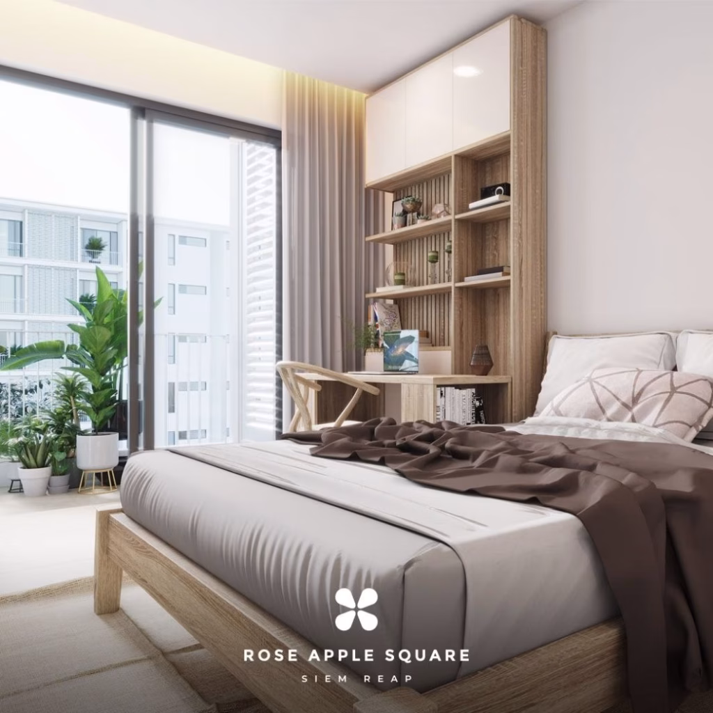 A modern bedroom at Rose Apple Square with a wooden bed, beige bedding, and brown throw blanket. A desk with shelves, books, and plants sits beside a large window framing natural light. Text reads Rose Apple Square Siem Reap.